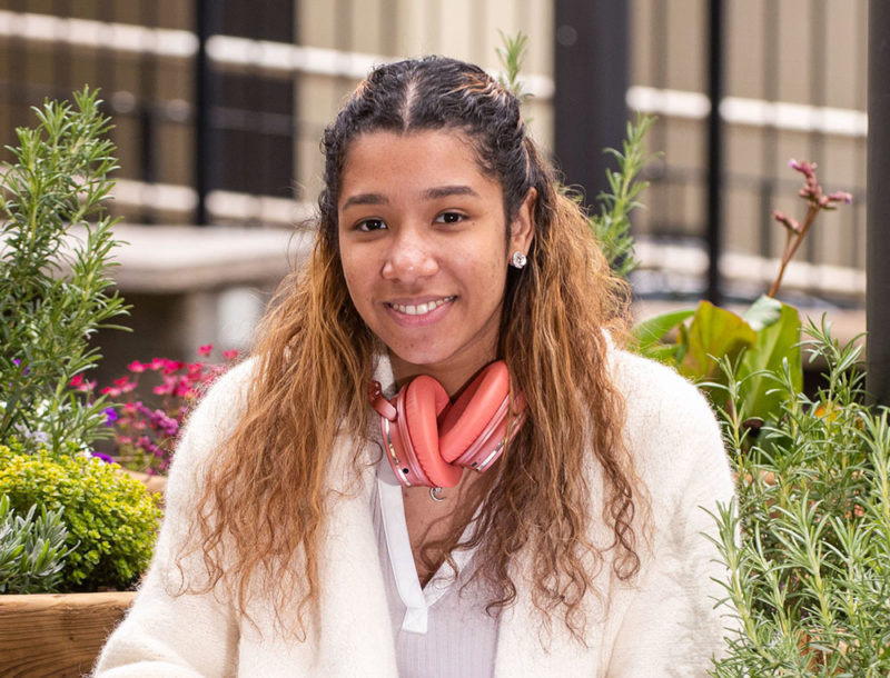 black female student wearing headphones sitting among flowers on campus smiling at camera