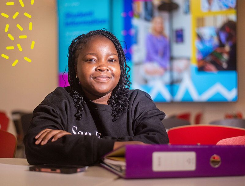 Smiling black female student sits at table with purple folder in front of digital screen