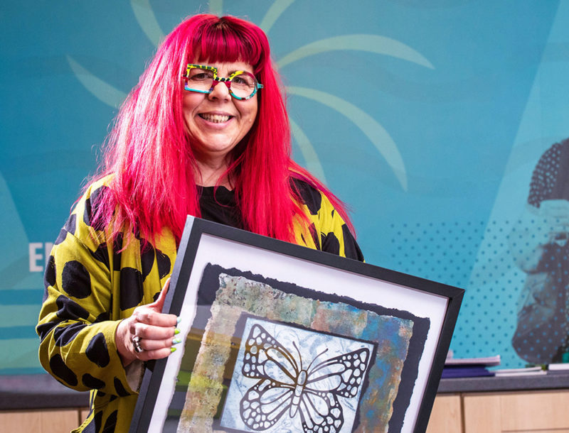 mature female art student with red hair holds her painting and smiles at camera