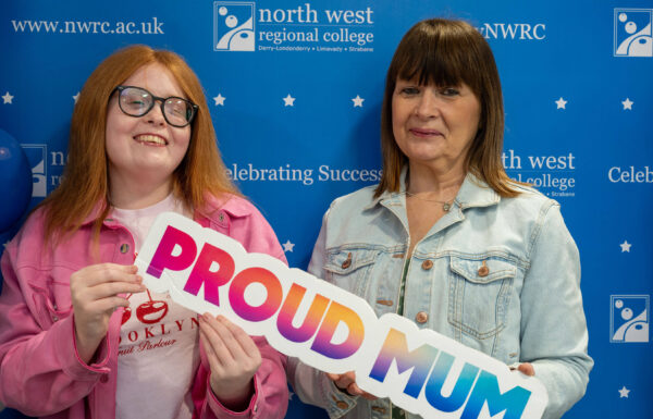 Two people at a college event holding a colourful “Proud Mum” sign, including one person with red hair and glasses.