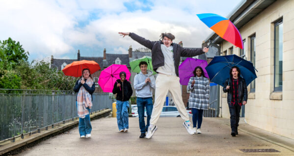 Young adults walking holding multi coloured umbrellas with one male jumping in the air