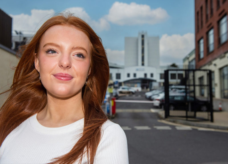 Aileen Barber, a female computing student with red hair smiling standing at the entrance to Strand Road Campus