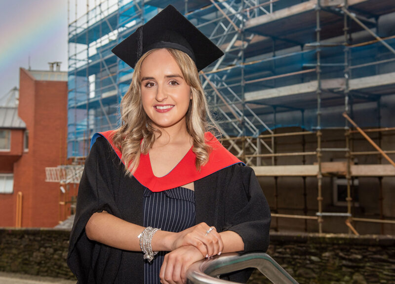 Female construction student Eva McLaughlin dressed in graduation gown and standing in front of scaffolding