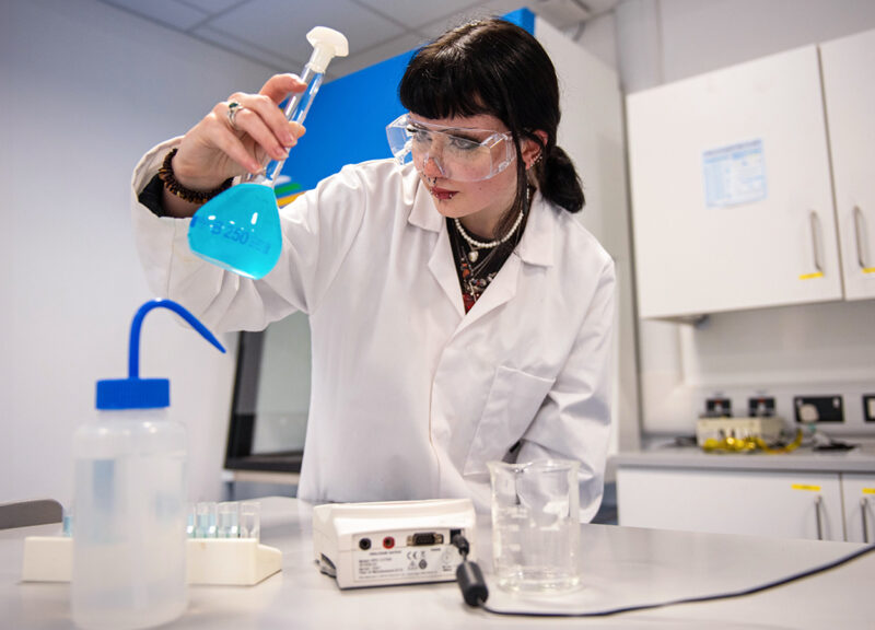 Female science student Hannah Kearney in white lab coat examining a beaker with blue fluid