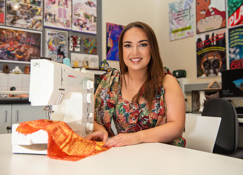 Art graduate Aoife Harvey sat at a table stitching orange fabric using a white sewing machine surrounded by student artwork on the walls