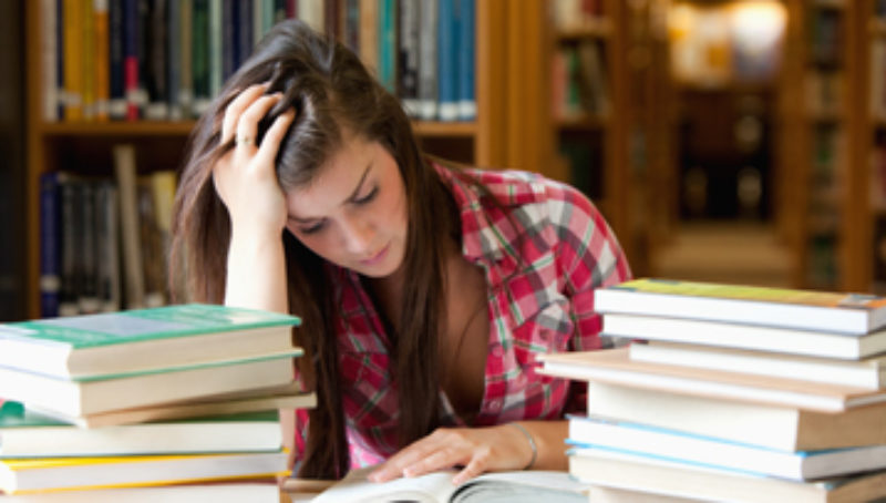 Female young adult student sitting at table in library reading a book, piles of books on table