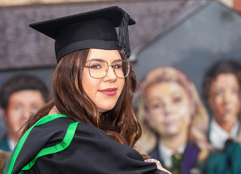 Female graduate smiling in front of the Derry Girls mural