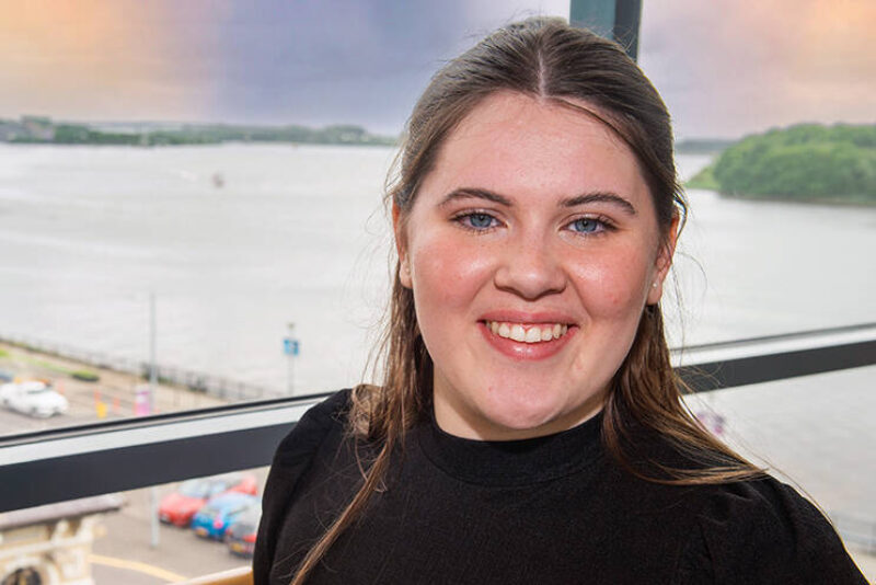 Female student smiling while standing in front of a window showing a view of the River Foyle