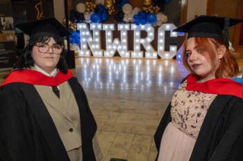 Two students in graduation gowns in front of light up letters that spell NWRC