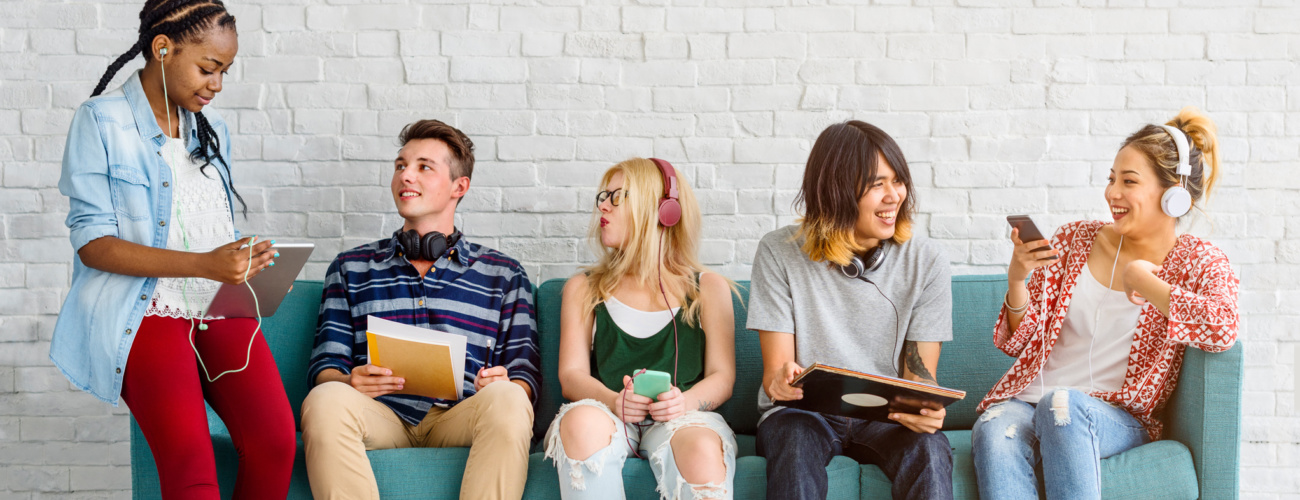 Group of young adult students sitting on sofa, using a variety smartphones, tablets and headphones.