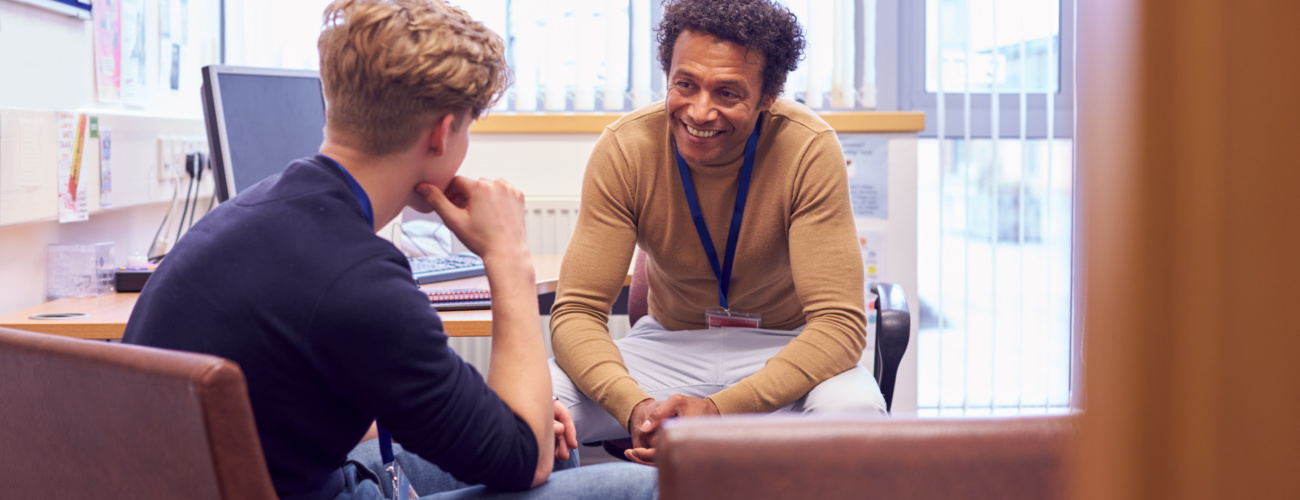 Staff member wearing lanyard in his office, chatting and smiling with student