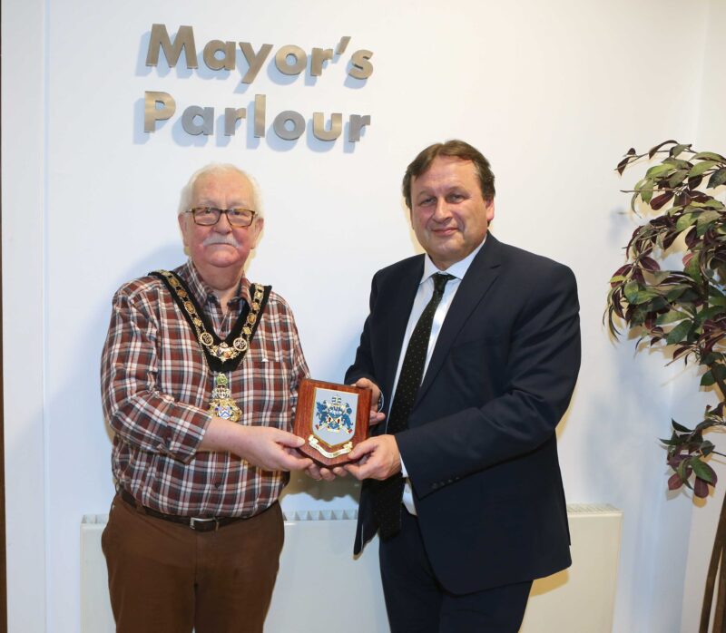 The Mayor, wearing the chain of office presents an award to another gentleman. The sign behind reads Mayor's Parlour. There is a plant on the far left.