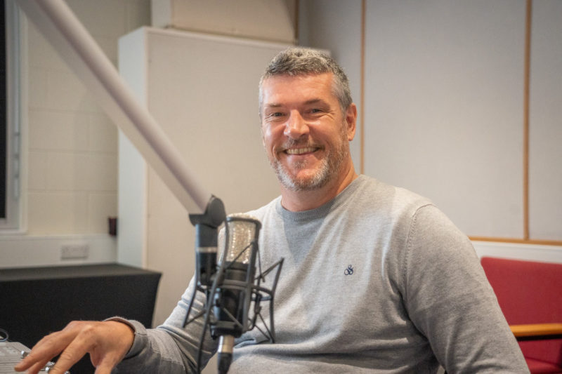 Man in grey jumper sitting in front of a radio mic.