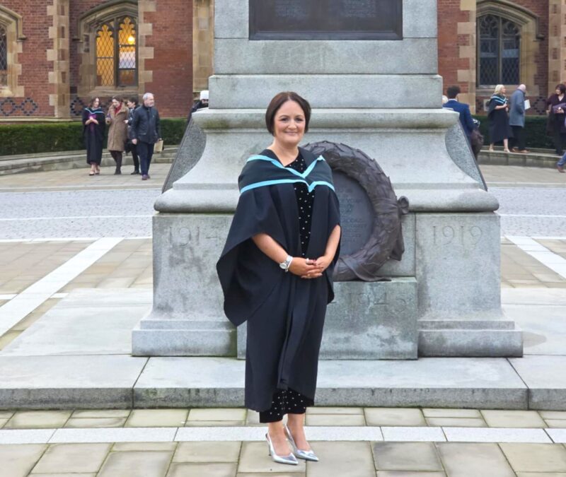 Lady in graduation robes stands outside a university building smiling