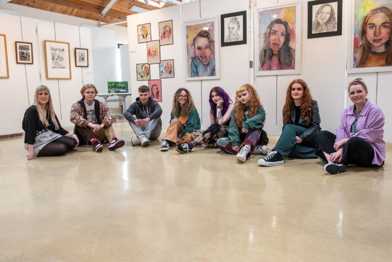 group of students sitting on the floor. Above the art is displayed on the walls.