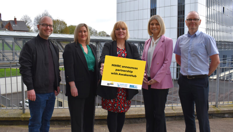 Two men and three women standing outside a college building. One of the women is holding a sign which states NWRC welcomes partnership with Awakenhub