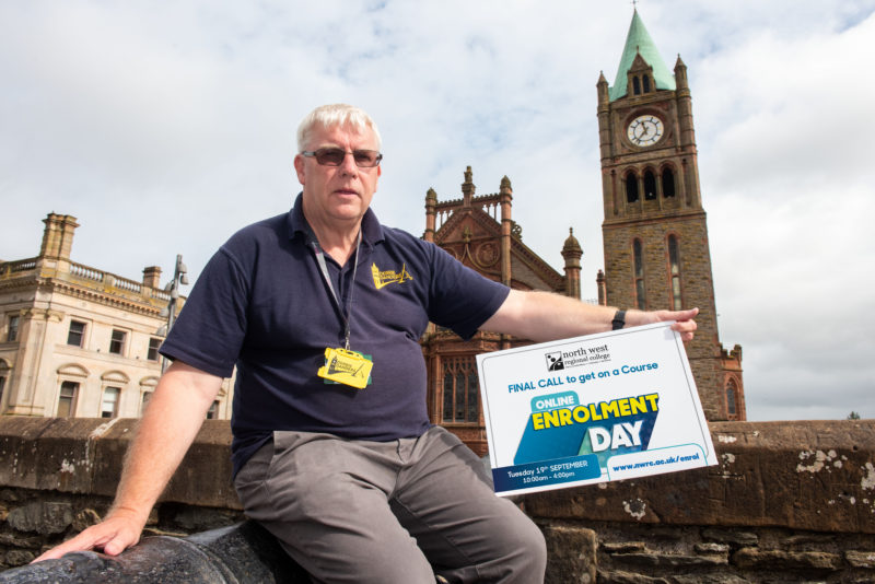 Man sitting on Derry's walls holding a sign about enrolment day. The guildhall is in the background,