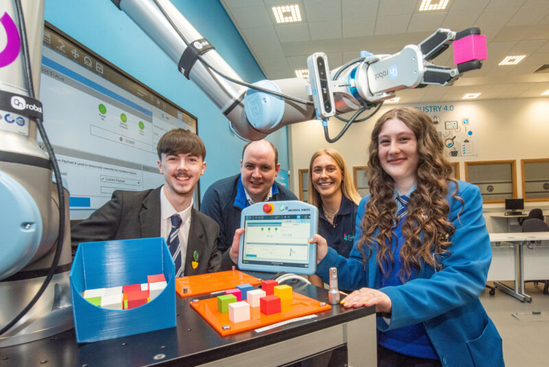 Two school pupils with a man and woman in a robotics room with equipment around them.