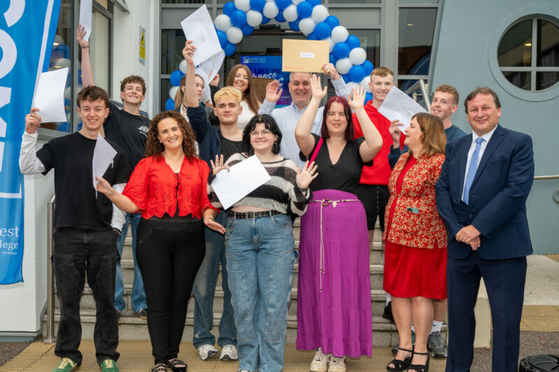 group of students with members of management outside a college holding up envelopes and cheering. There is a balloon arch and flags.