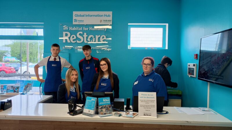 Six people standing behind the till in a charity shop called Restore - They are wearing blue aprons.