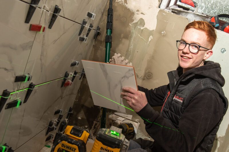 Student with glasses pictured in a house where he is tiling a bathroom