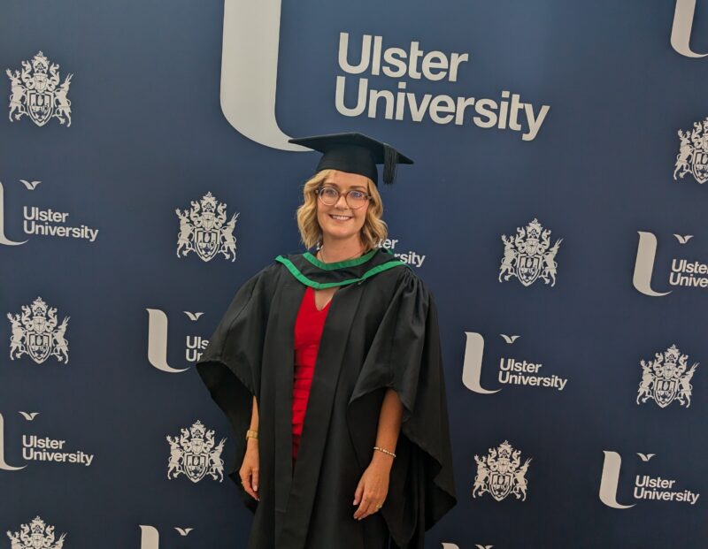 Lady with blonde hair and glasses wearing a graduation hat and gown with Ulster University branding