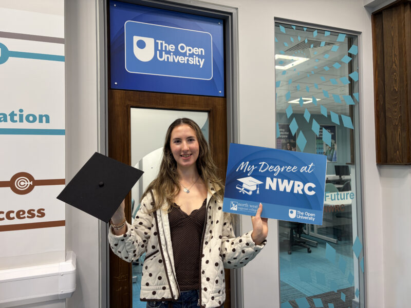 Student in a college room holding a sign that says My Degree at NWRC, and a black mortar board. A sign with the Open University logo is behind her