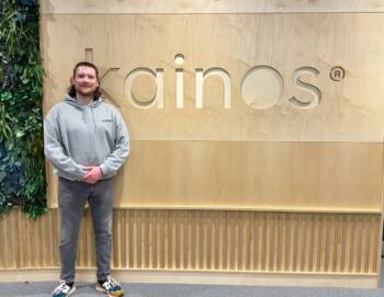 Man dressed in grey hoodie with long hair in front of a wooden sign spelling out Kainos.