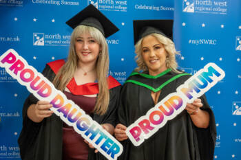 Two women at graduation holding signs that say proud mum and proud daughter