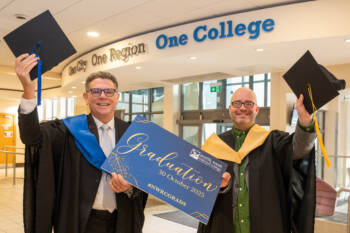 Two people in graduation robes hold a blue sign that states Graduation - October 30