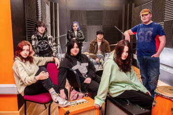 Seven students in a music studio surrounded by drums, speakers and microphones