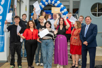 group of students with members of management outside a college holding up envelopes and cheering. There is a balloon arch and flags.
