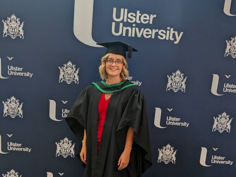 Lady with blonde hair and glasses wearing a graduation hat and gown with Ulster University branding